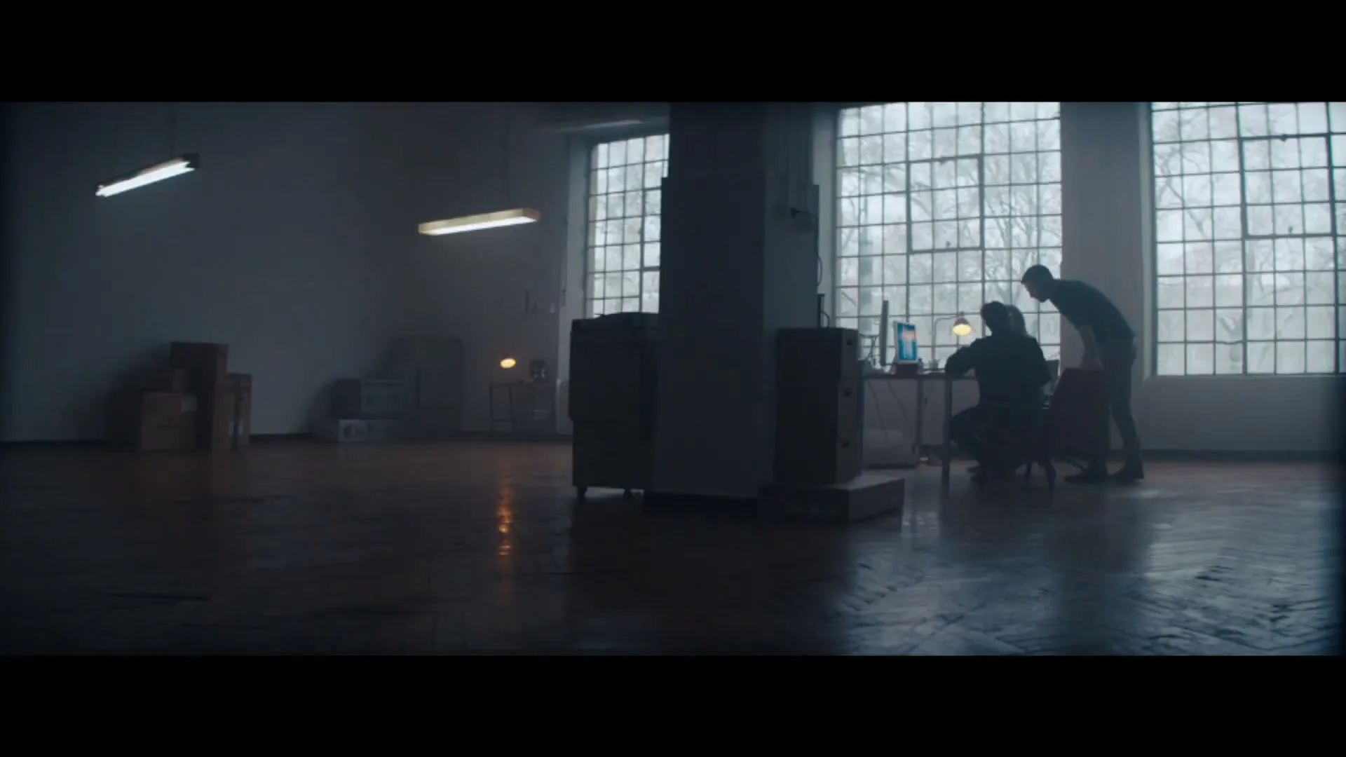 Two men are working on computers in a dimly lit, empty industrial loft with large windows looking out onto bare trees.