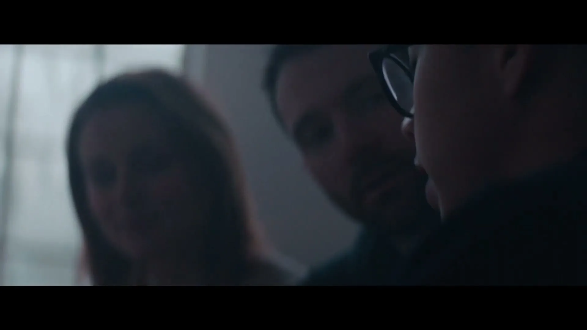 Three people, two men and one woman, are seen in a dark, intimate setting, with one man wearing glasses in the foreground, looking down.