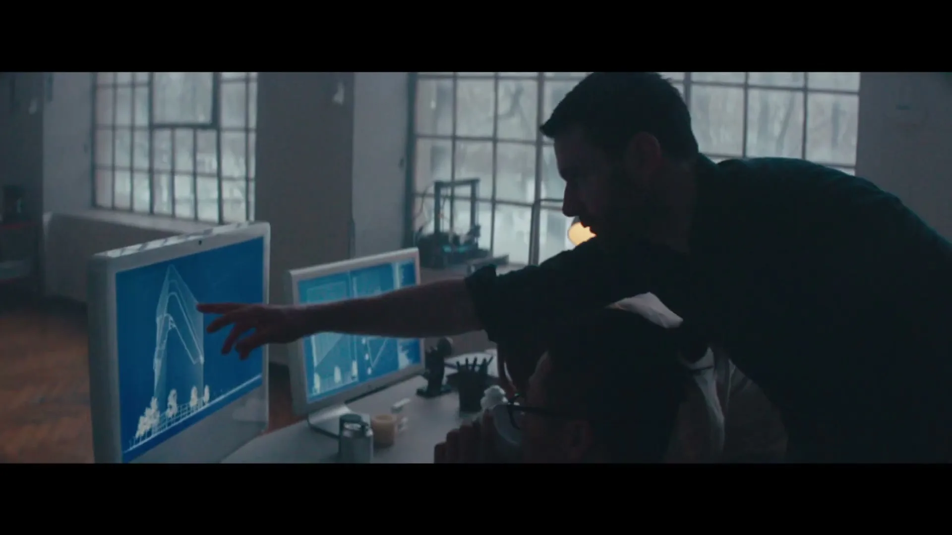 Two men in an industrial office space look at computer screens displaying architectural blueprints, with one man pointing at a design.