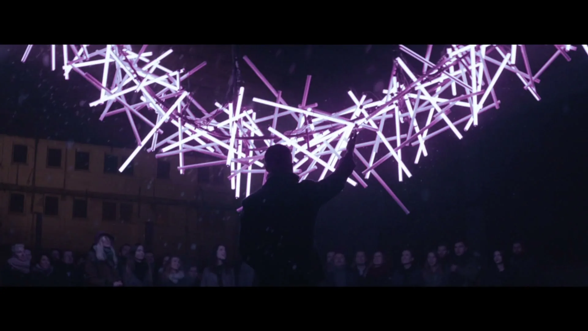 A silhouetted man stands in a crowd, looking up at a large, glowing purple light sculpture in the night sky, with a building in the background.