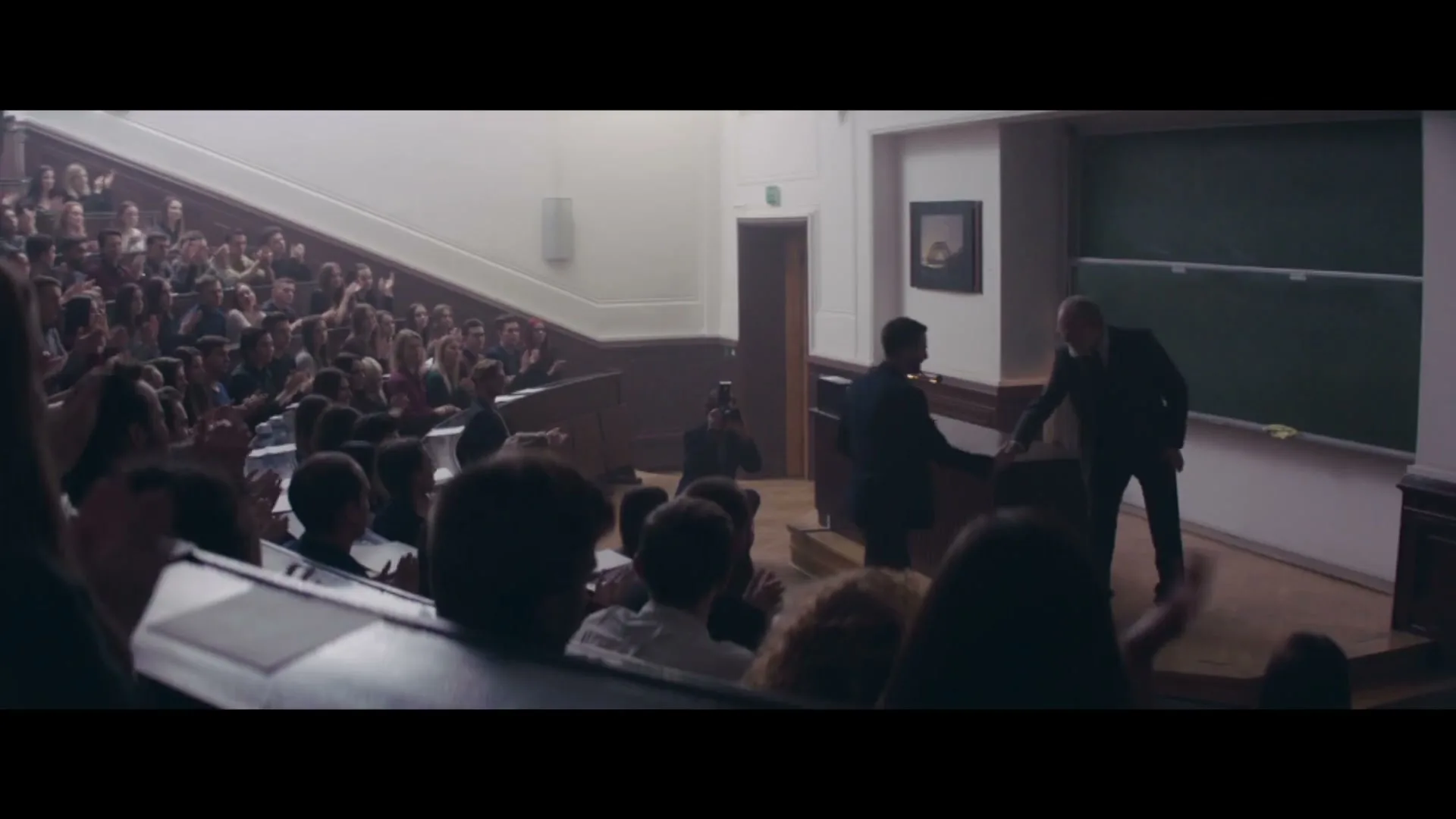 A young man in a suit shakes hands with an older man in a suit at the front of a lecture hall, as a large audience applauds.