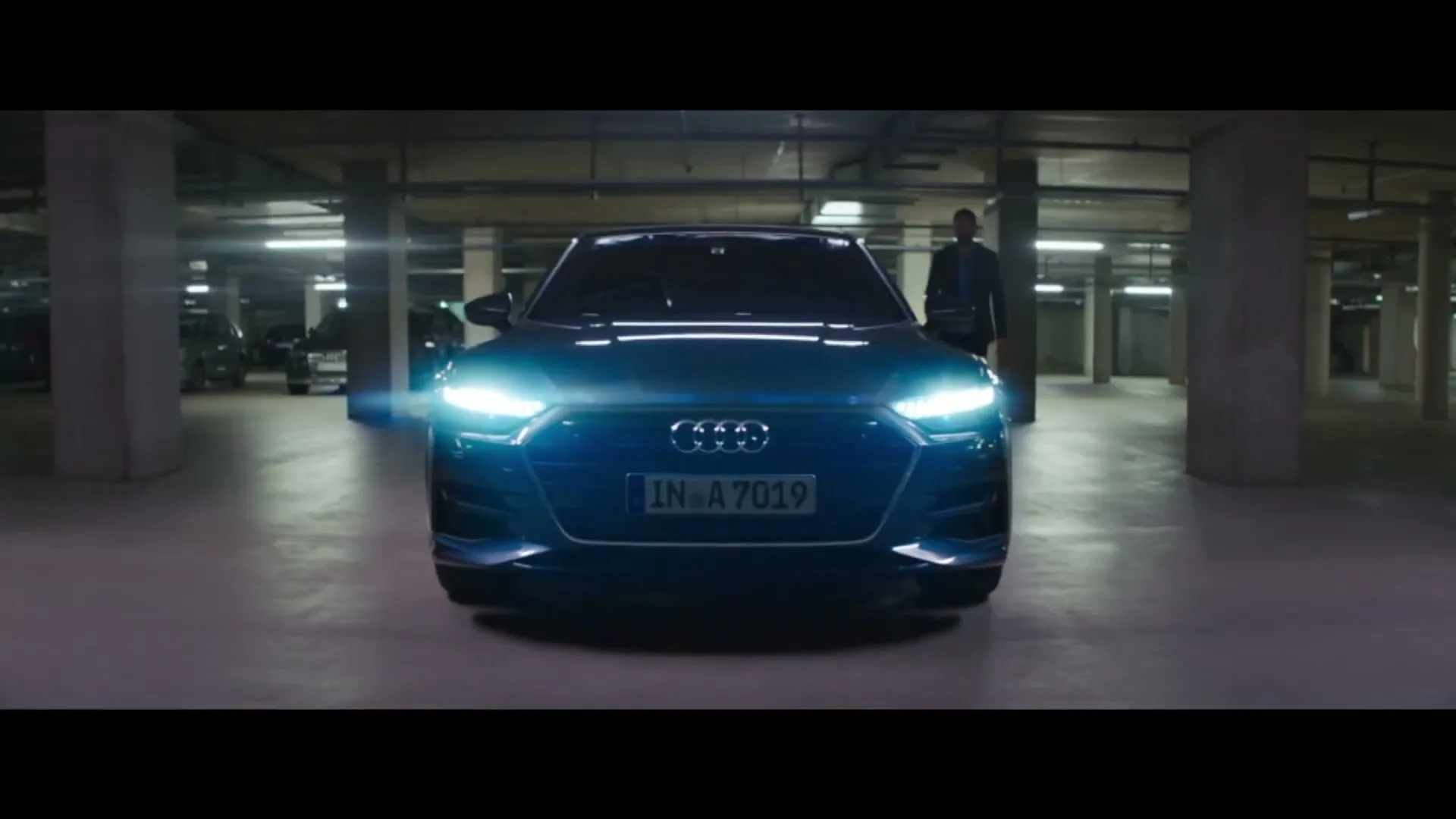 A dark blue Audi car with bright headlights is parked in an underground parking garage, with a man standing behind it.