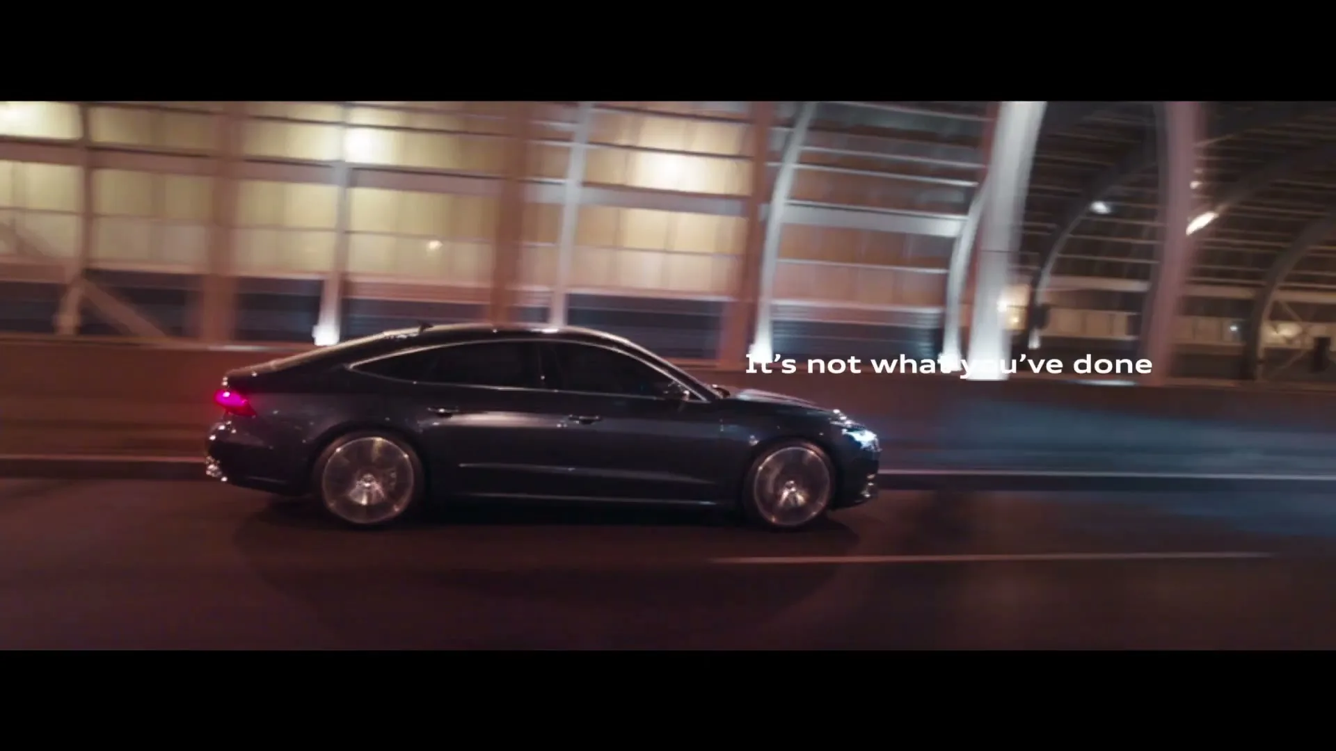 A dark grey Audi car drives at night on a highway with blurred lights in the background, with white text appearing on screen.