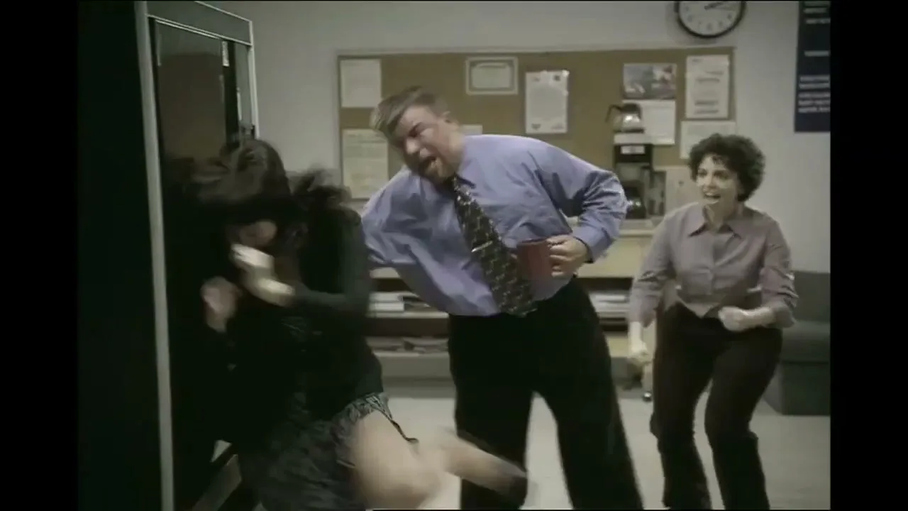 A man and two women in an office are shaking a vending machine to get a snack, with one woman hitting the machine and the others cheering her on.