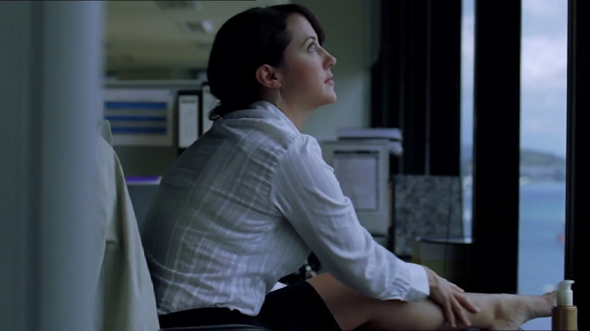 A woman in an office chair applies lotion to her leg while looking out a window at a distant ocean view.