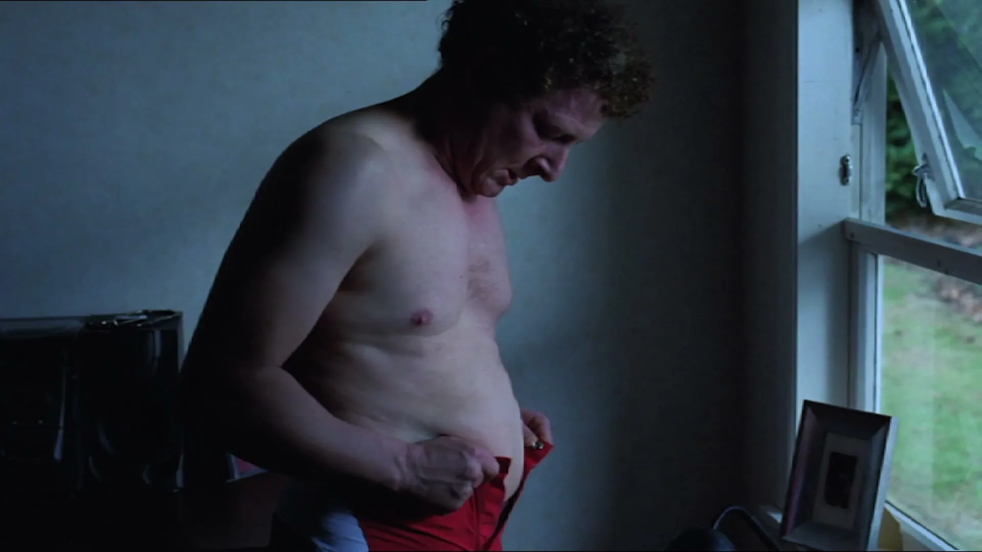 A middle-aged man with curly hair, shirtless, struggles to button his red shorts while looking down at his stomach in a dimly lit room.