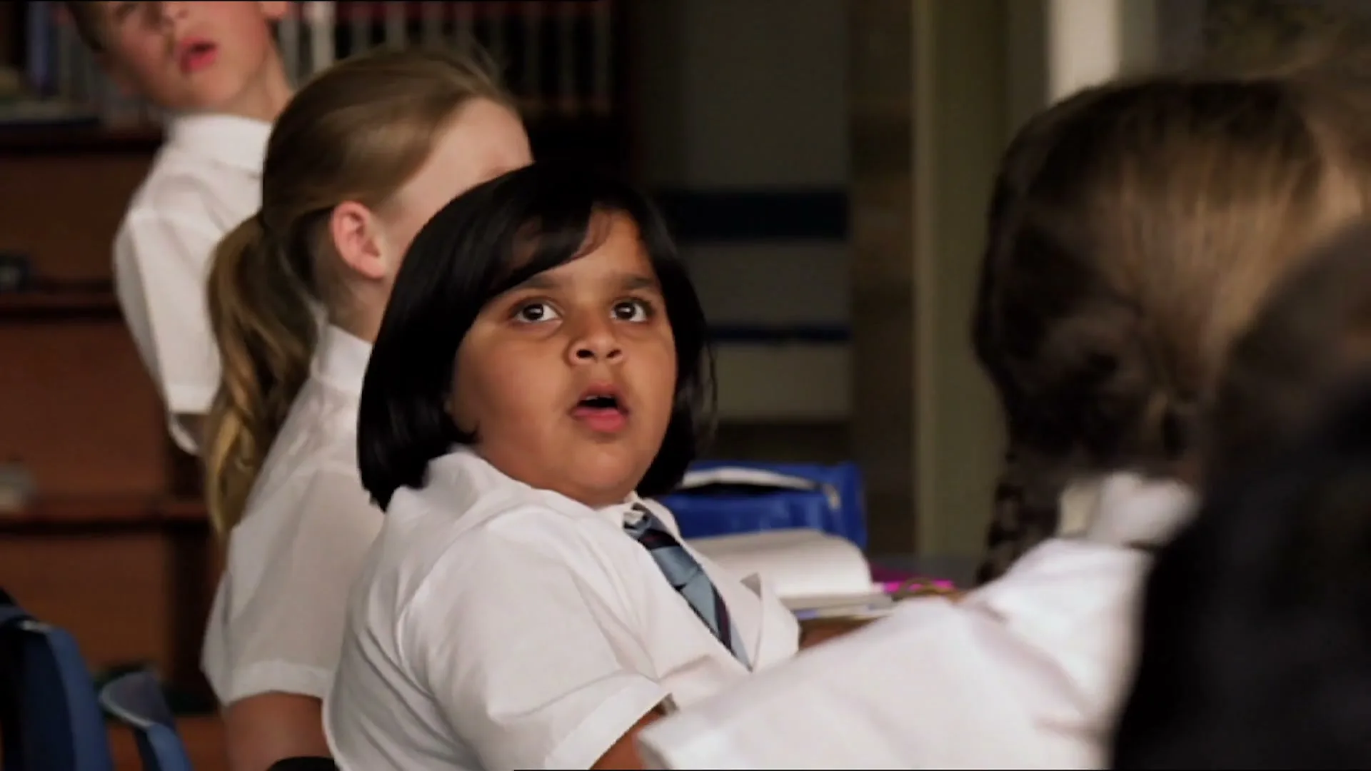 A young girl with dark hair and a white school uniform looks up with a surprised expression in a classroom.