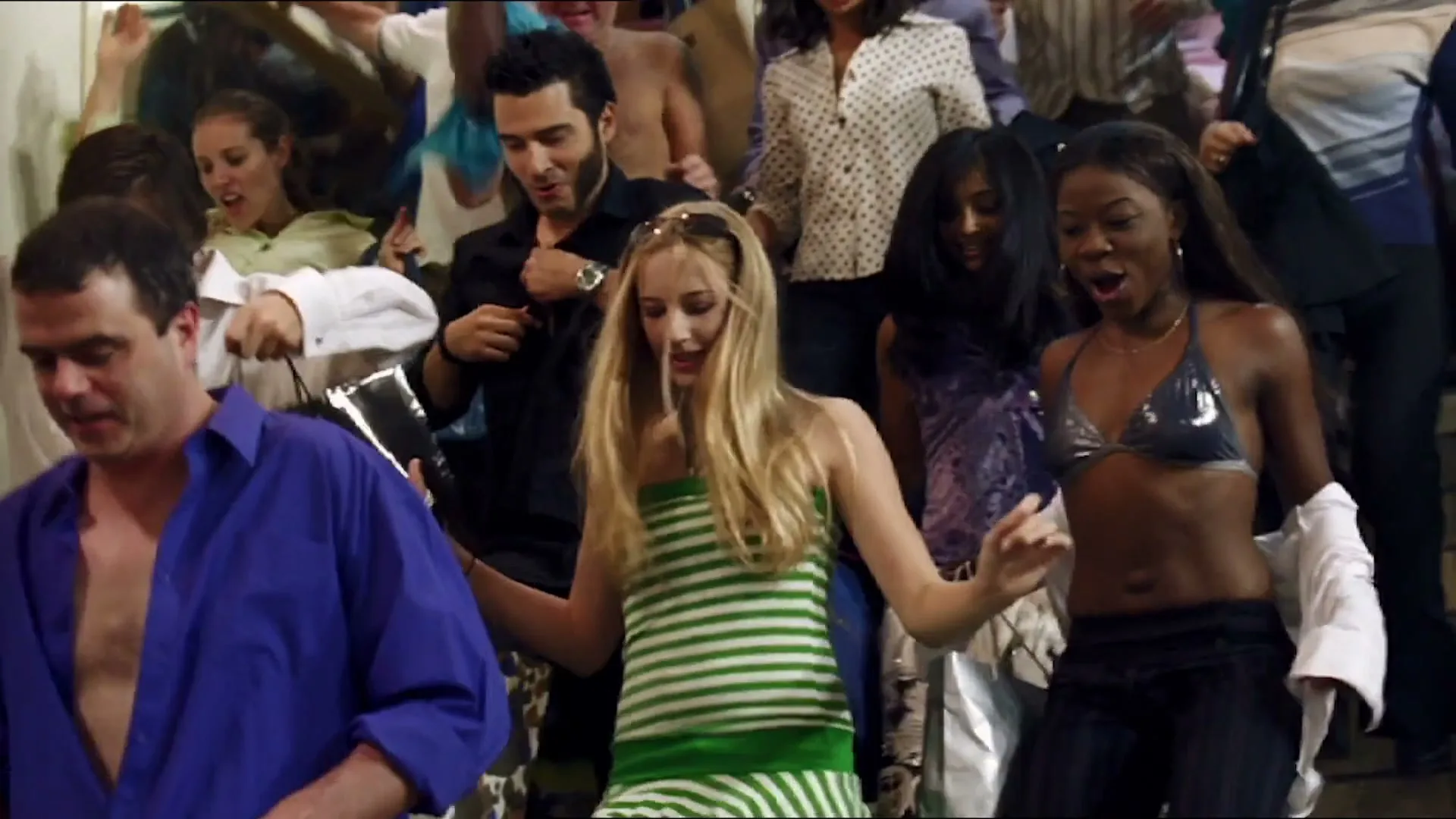 A diverse group of people, dressed in various casual outfits, dance energetically and joyfully in a crowded indoor space, possibly a store or club.
