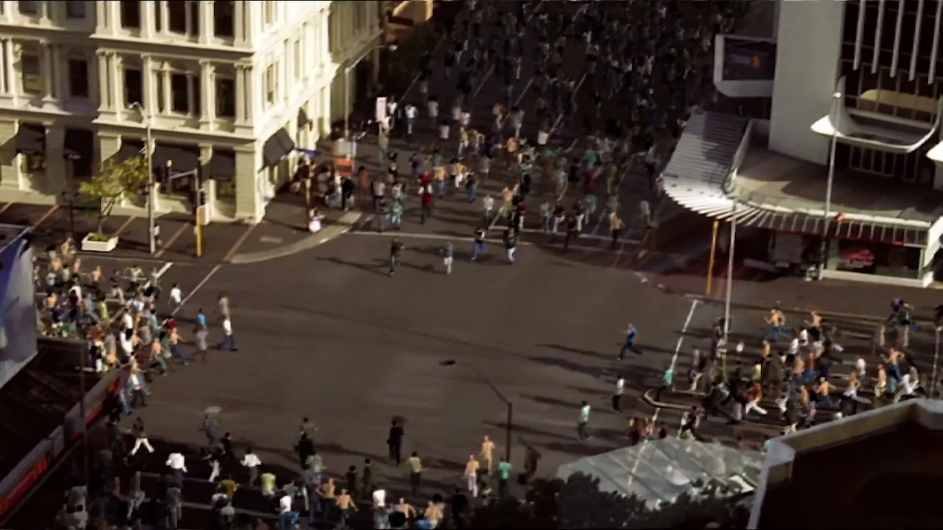 An aerial shot shows a busy city intersection with hundreds of people running across the streets and sidewalks, surrounded by tall buildings.