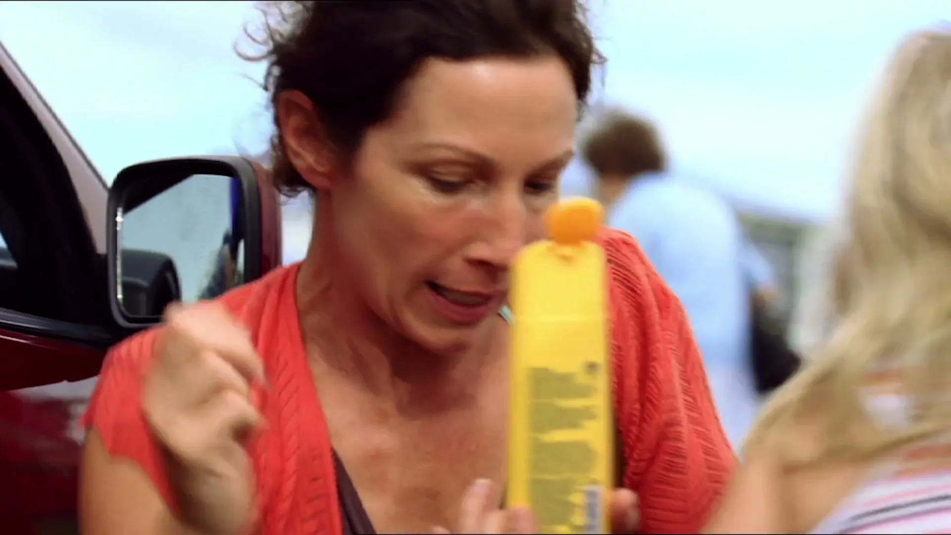 A mother with dark hair, wearing a coral cardigan, applies sunscreen to her blonde-haired child near a red car at a beach.