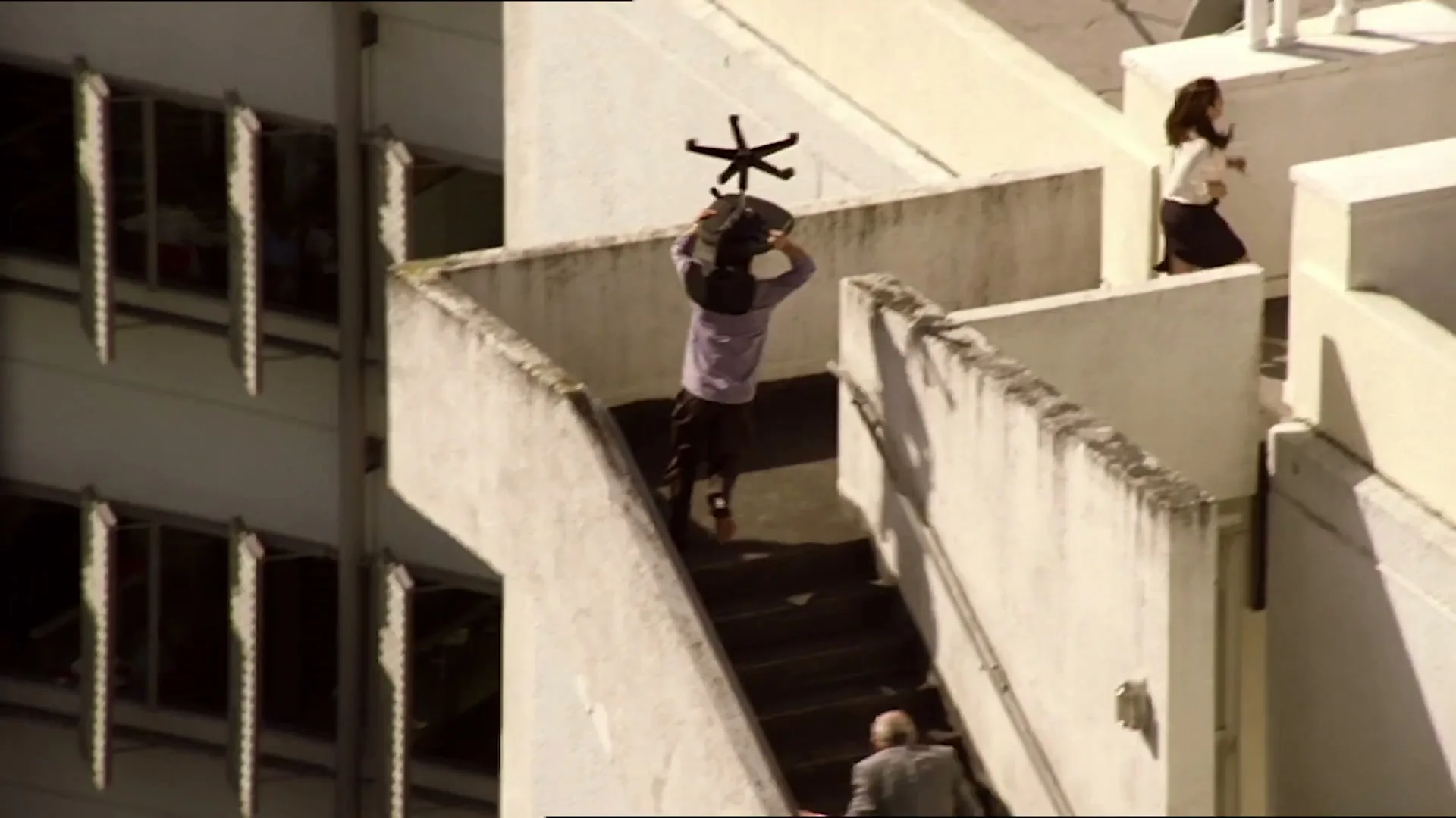 A man carries an office chair on his shoulders, running up an outdoor staircase, followed by other people, in a high-angle shot.