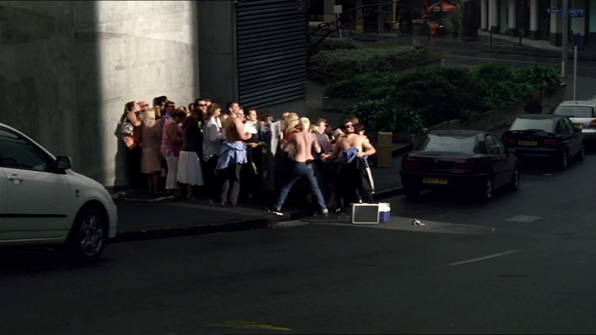 A diverse group of people, some shirtless, are gathered on a sunlit city sidewalk next to parked cars, appearing to be waiting or observing something.