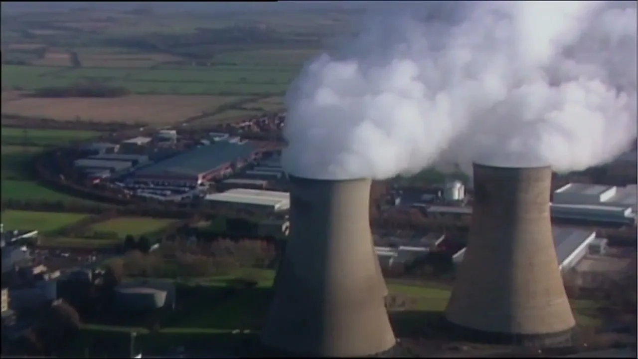 An aerial view shows two large cooling towers emitting white smoke into the sky, with industrial buildings and green fields in the background.