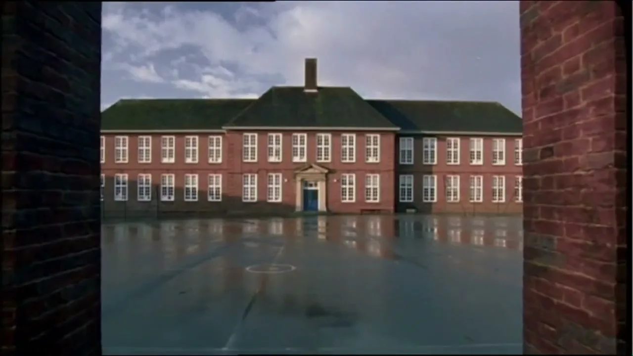 A wide shot of a red brick school building with many windows, seen from across a wet, empty playground, framed by brick walls on either side.