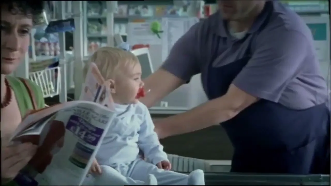 A woman in a green top and red necklace reads a magazine while a baby with a pacifier sits on a counter, then a man in a purple shirt picks up the baby in a store.