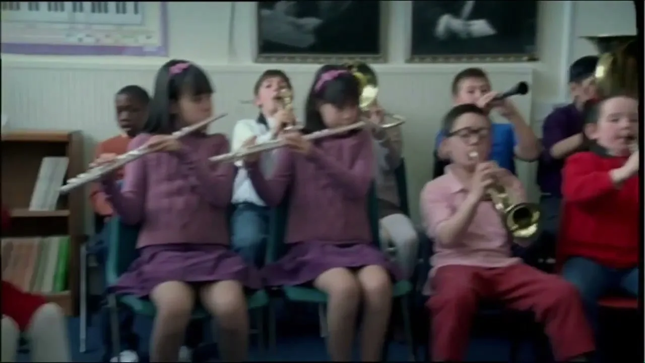 A diverse group of children in a school band play various instruments, some struggling, while an older male conductor with long hair directs them in a classroom.