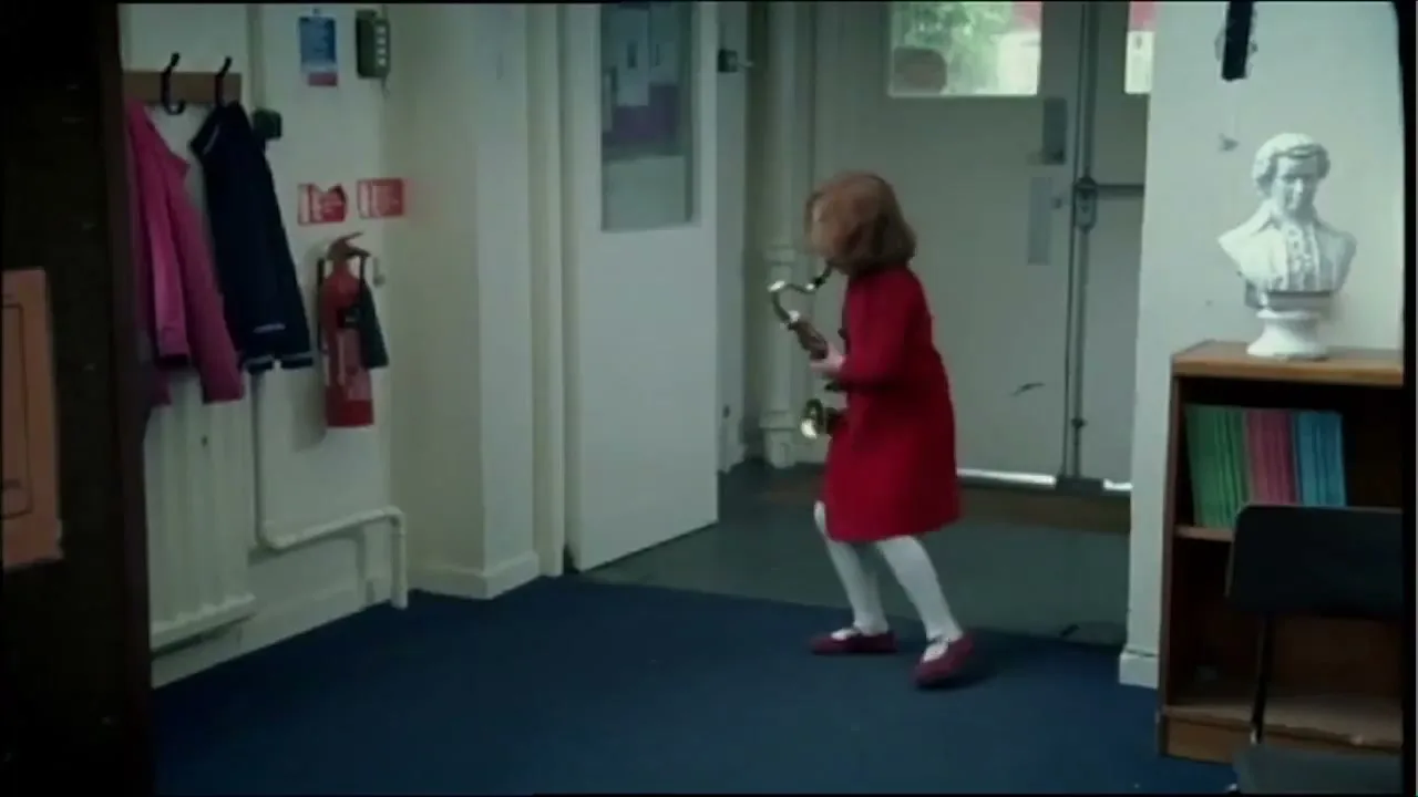 A male conductor with long grey hair leads a children's orchestra in a classroom, then a young girl in a red dress runs out of the room with a saxophone.