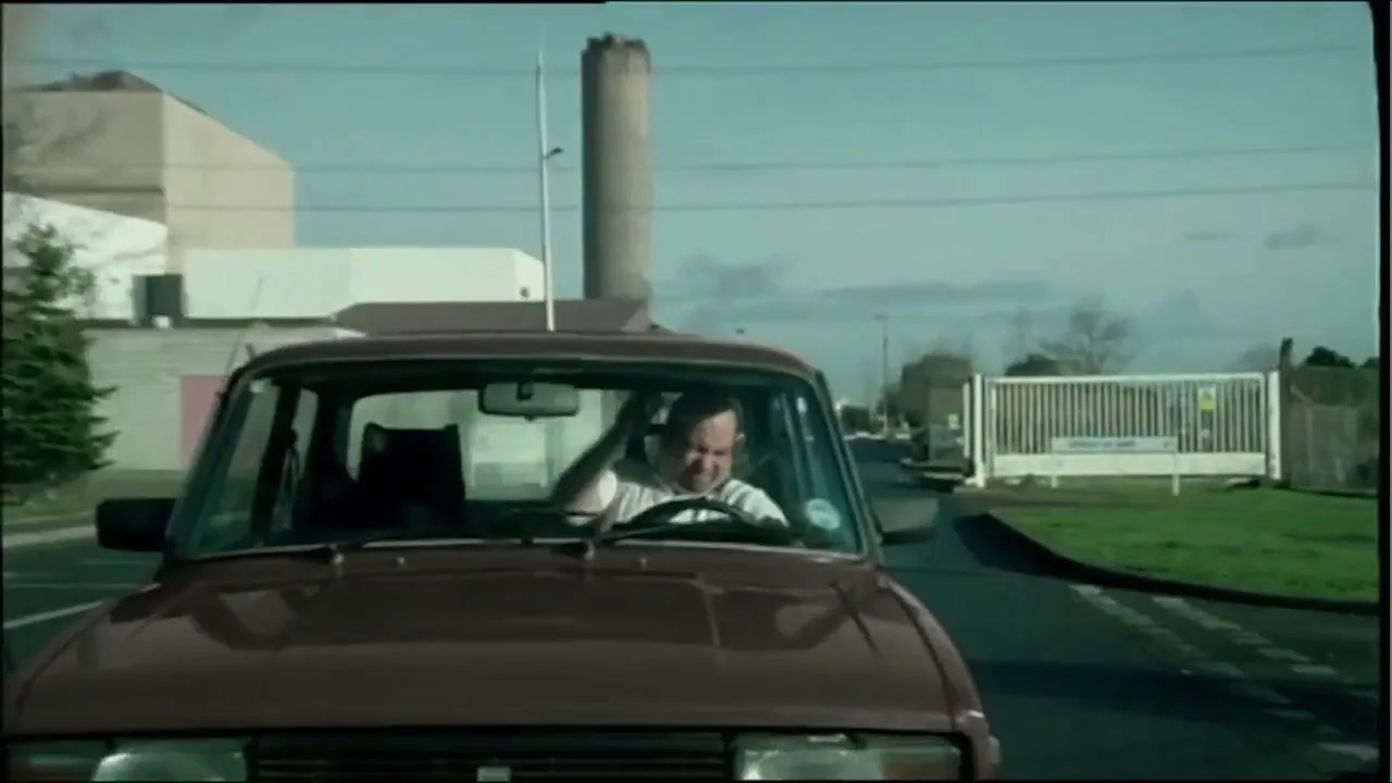 A man in a white t-shirt drives a brown car past a power plant, looking stressed and scratching his head.