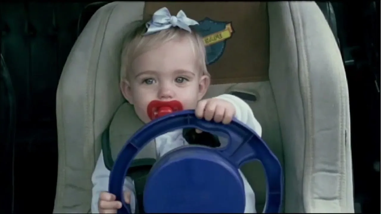 A baby girl with a pacifier and a blue toy steering wheel sits in a car seat, looking directly at the camera.