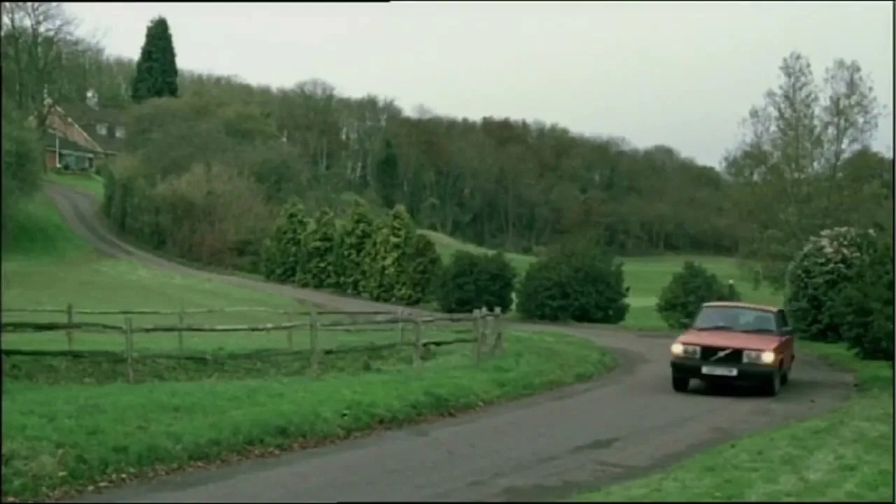 A red car drives along a winding road through a green, hilly landscape with trees and a house in the distance under an overcast sky.
