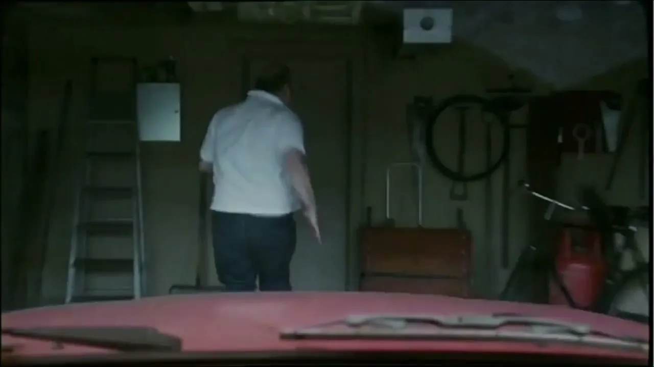 A bald, overweight man in a white polo shirt and jeans runs towards a door in a cluttered garage, viewed from inside a car.