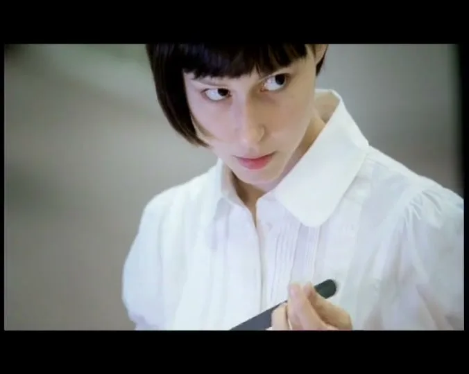 A young woman with a bob haircut and white shirt looks intently at a nail file she holds, in an interior setting.