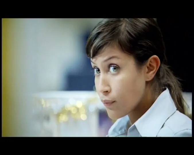 A young woman with dark hair tied back, wearing a light blue collared shirt, looks intently at something off-screen in an interior setting.
