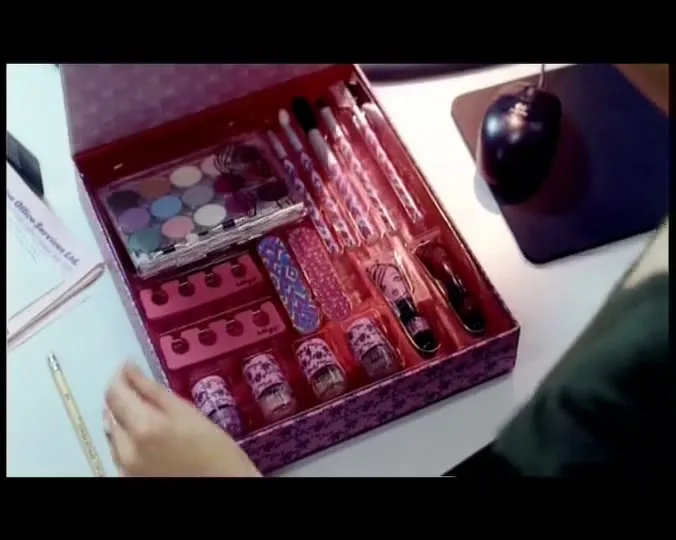 A woman's hands open a makeup kit designed to look like a magazine, revealing various cosmetics and tools inside, on a desk.