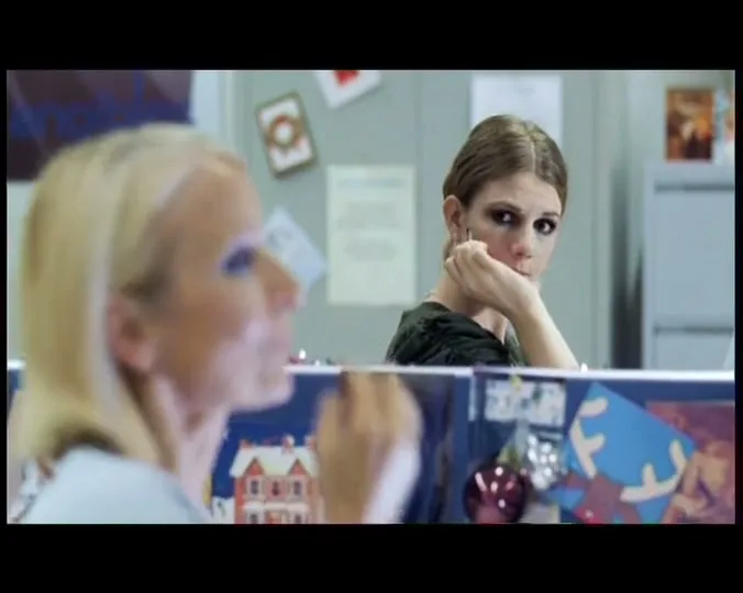 Two women in an office cubicle apply makeup; one looks bored while the other smiles with closed eyes, enjoying the moment.