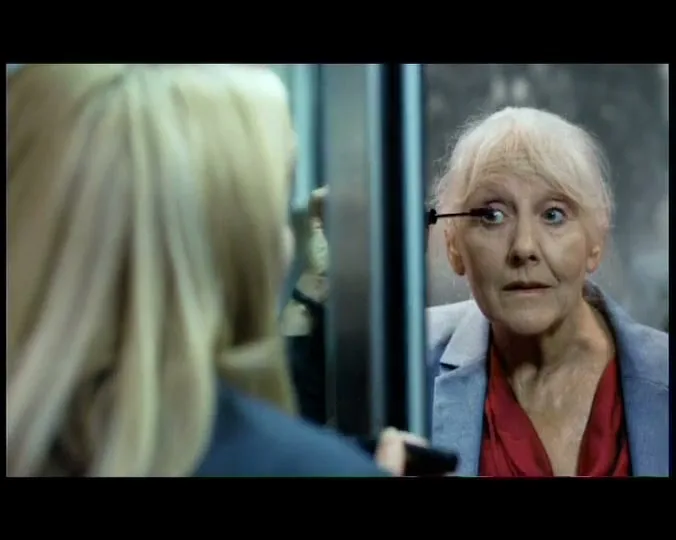 A blonde woman applies mascara in a mirror, transforming into an older woman with white hair, in an elevator.