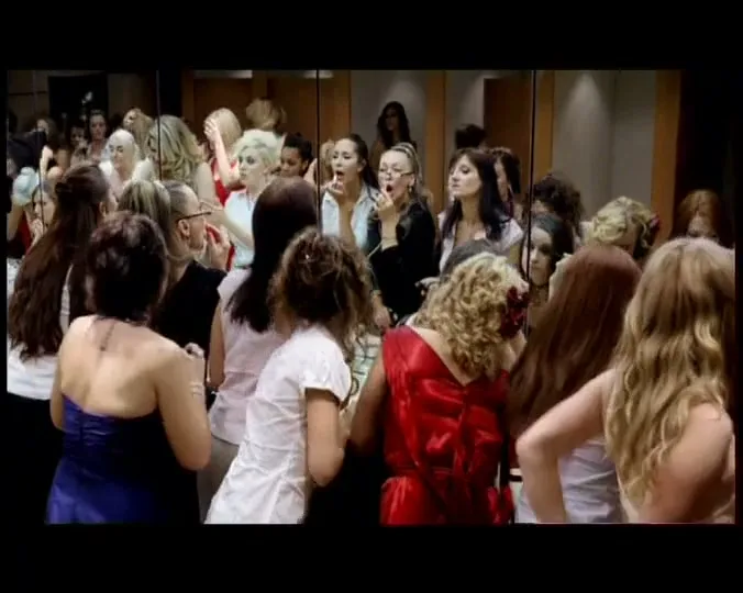A diverse group of women, dressed in various outfits, apply makeup and adjust their hair in front of a large mirror in a changing room.