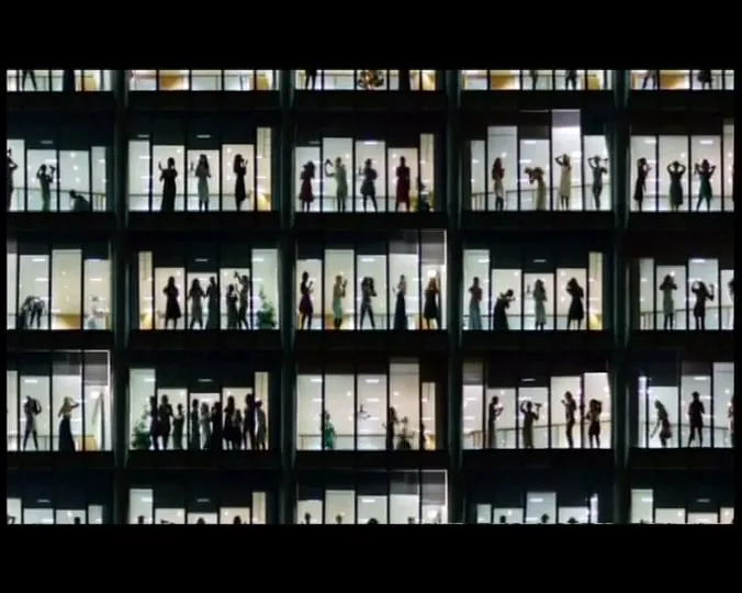 Silhouetted women in various poses are visible through the illuminated windows of a large office building at night.