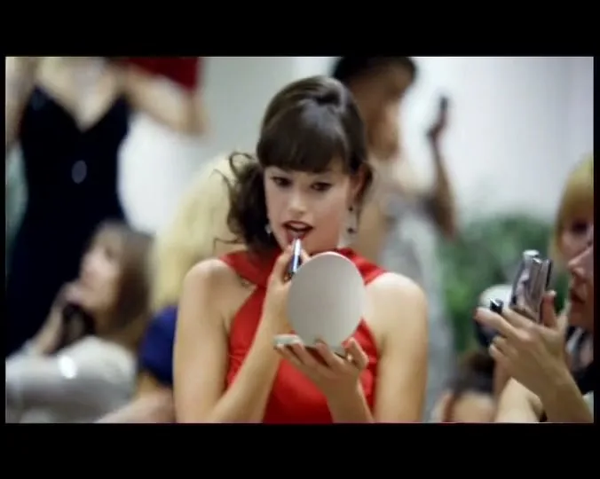 A young woman in a red dress applies lipstick while looking into a compact mirror, surrounded by other women getting ready in a bustling room.