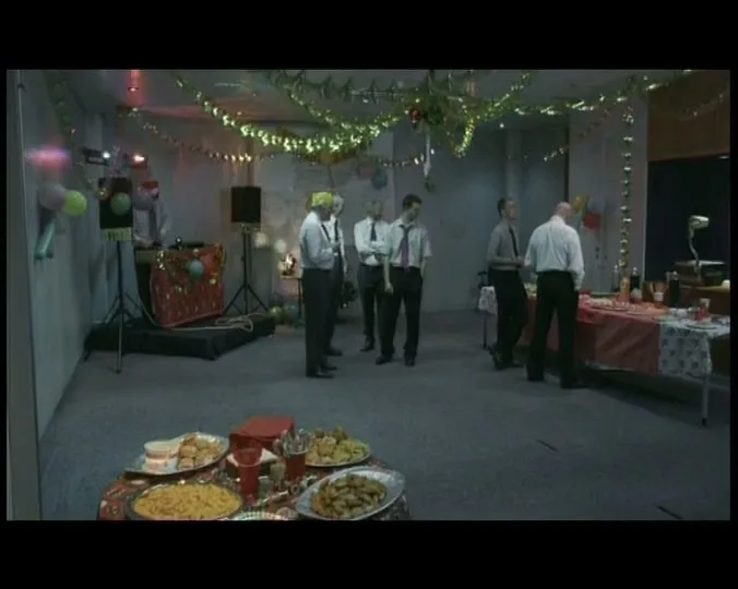 A group of male office workers in shirts and ties stand awkwardly at a sparsely attended Christmas party in an office.
