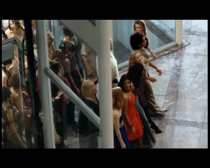 A diverse group of women in various outfits walk down a modern, brightly lit shopping mall corridor, some smiling and looking around.
