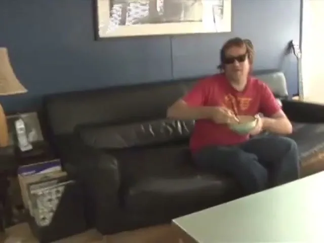 A man in sunglasses and a green shirt looks at the camera, then takes off his sunglasses and throws them to another man on a couch, who catches them and smiles.