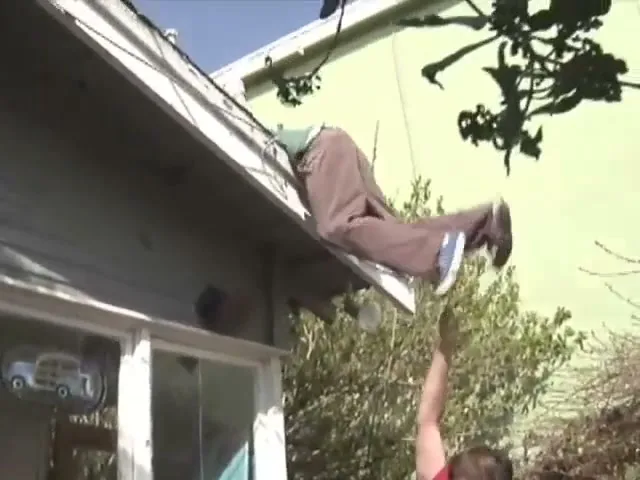 A young man in a red shirt helps another young man in a green shirt and brown pants climb onto a house roof in a sunny backyard.