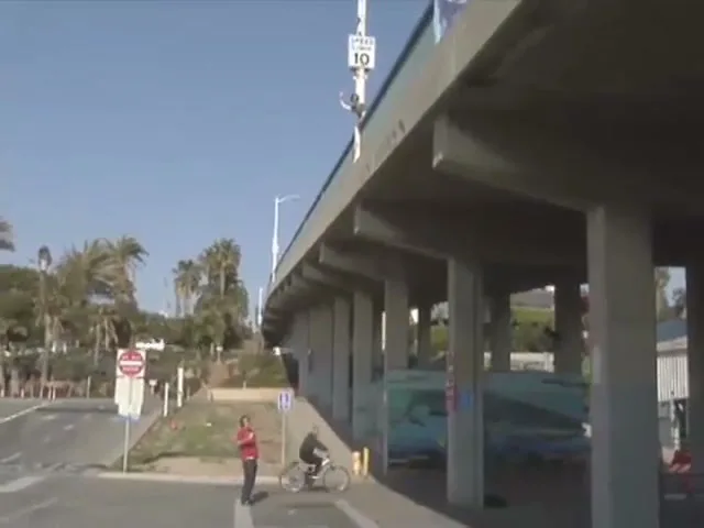 A man in a red shirt stands on a sidewalk next to a highway overpass as a cyclist rides past, with palm trees in the background.