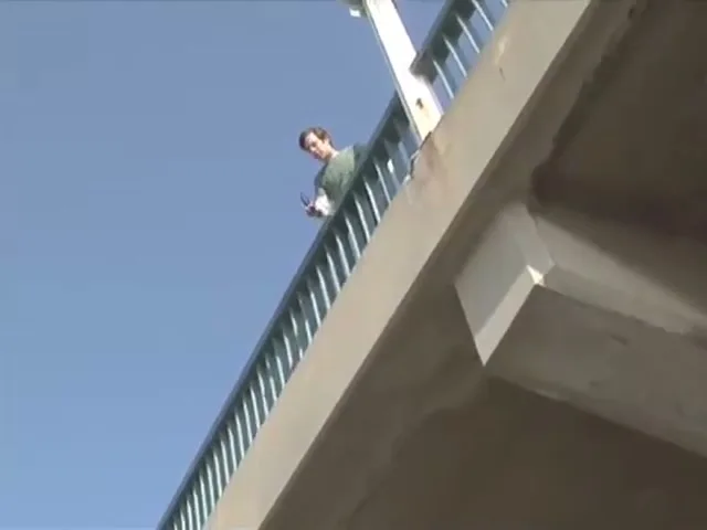 A man in a green shirt stands on a bridge, taking off his sunglasses and dropping them, as the camera looks up from below.