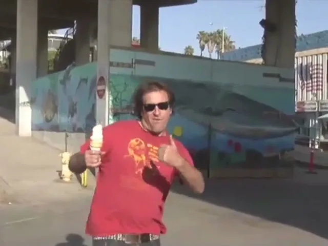 A man in a red shirt and sunglasses holds an ice cream cone and points to himself under a bridge with a whale mural.