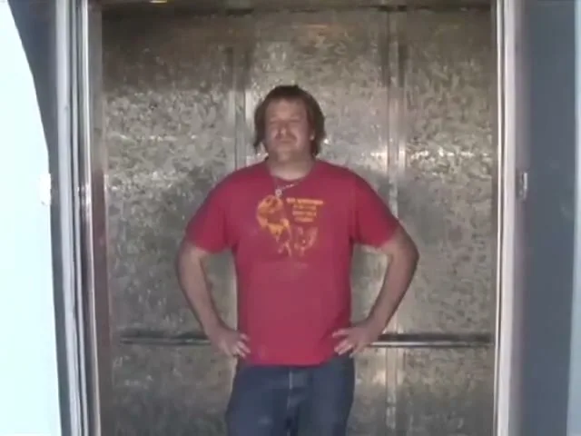 A man in a red t-shirt and jeans stands in an elevator, looking at the camera, as the doors close on him.