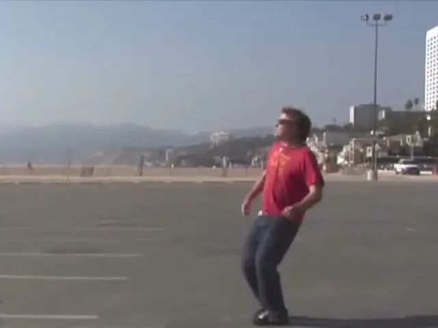 A young man in a green shirt and brown pants attempts to kick up a skateboard, then a man in a red shirt and jeans catches sunglasses in a parking lot by the beach.