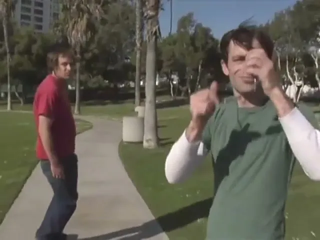 Two young men are skateboarding in a park on a sunny day, one throws sunglasses to the other who catches them and puts them on.