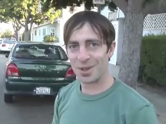 A man with brown hair and a green shirt smiles and looks at the camera on a suburban street with cars parked in the background.