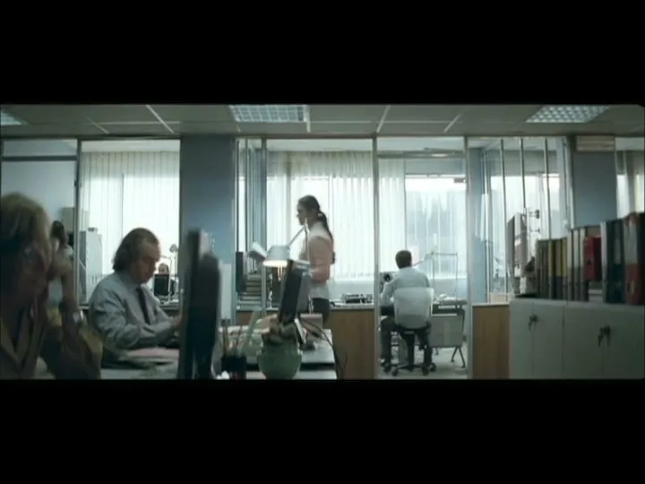 A wide shot of an office shows several people working at their desks, with a woman in a pink top walking through the middle.