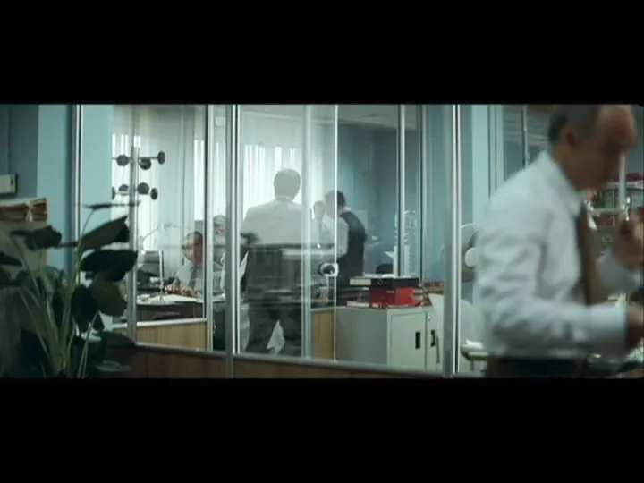 A male office worker in a white shirt and tie walks past the camera, while other workers are visible through glass partitions in a busy office.