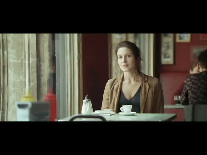 A woman with dark hair in a brown jacket sits at a table in a diner, looking directly at the camera with a slight smile.