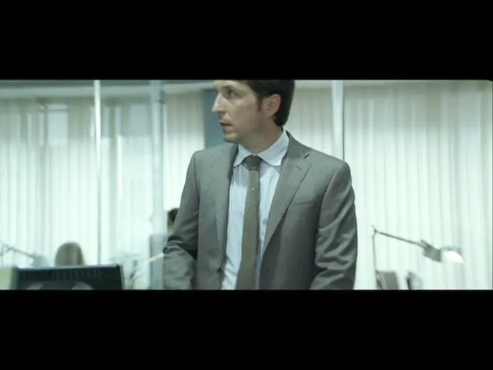 A man in a suit looks around nervously in a modern office with cubicles and blinds.