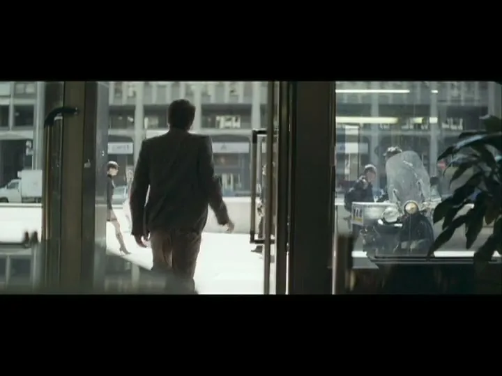 A man in a suit walks through glass doors, leaving a building and entering a busy city street with people and a motorcycle.