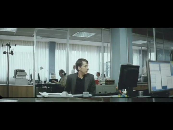 A man in a suit jacket sits at a desk in a modern office, looking around with a surprised expression.